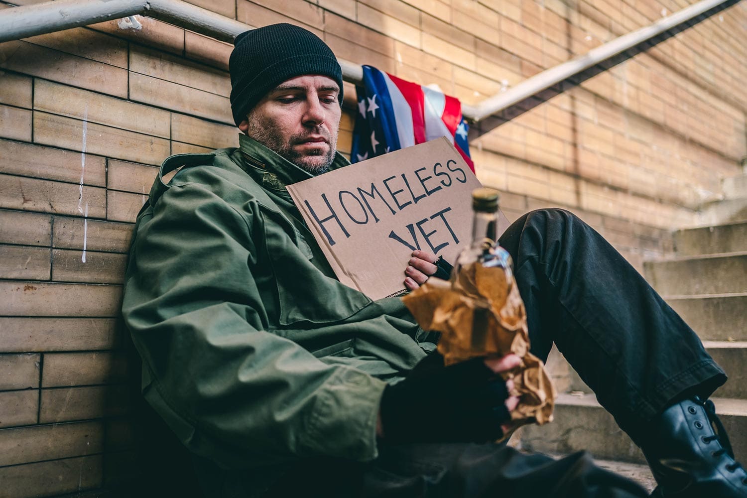 Featured image for VA Homeless Program, showing a homeless Veteran sitting on the stairs with a bottle of alcohol in one hand and holding up a sign in his other hand